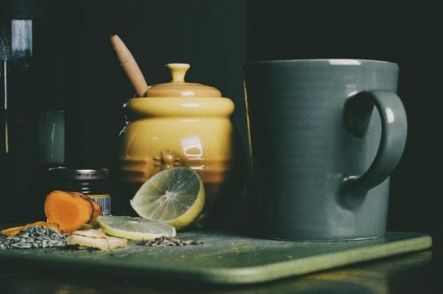 A ceramic cup and ceramic pot next to slices of lemon, turmeric, and seeds.