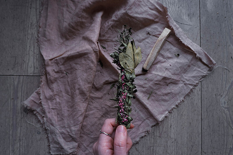A hand holding a smudge stick with shamanic herbs and small purple flowers.
